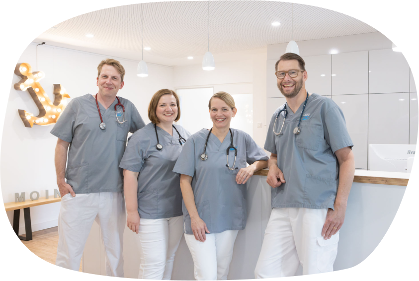 Vier lächelnde Ärztinnen und Ärzte in grauen Kasacks und weißen Hosen mit Stethoskopen um den Hals stehen gemeinsam am Empfangstresen der Gemeinschaftspraxis-Husum